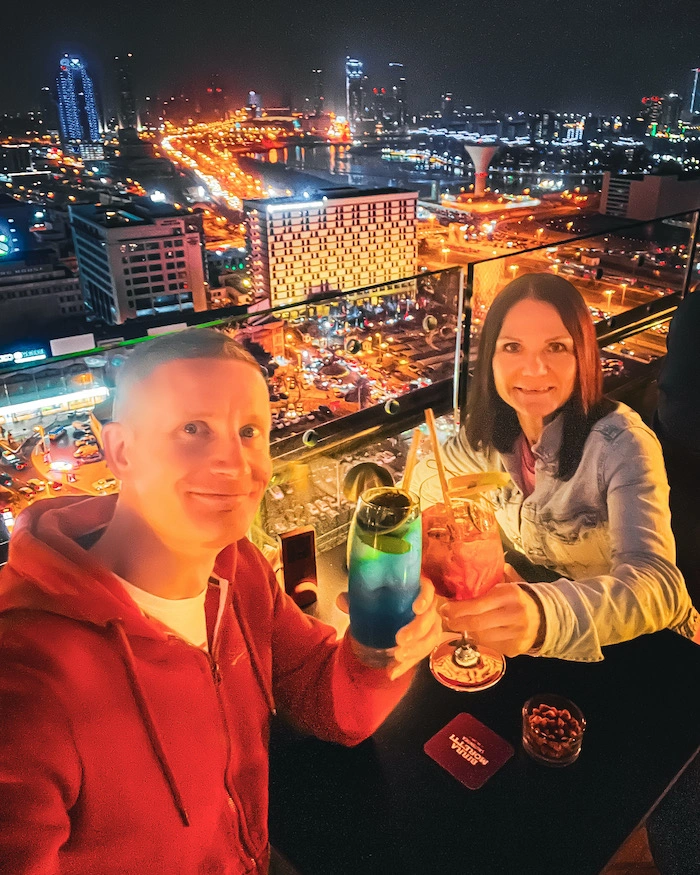 parsikunta drinkeillä pilvenpiirtäjän baarissa, taustalla näkyy iltavalaistu Manama City.